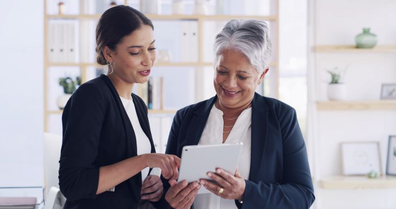 Dos mujeres con vestimenta profesional, en una oficina luminosa, revisan una tableta; la mujer más joven señala la pantalla mientras la otra, de pelo canoso, sonríe.