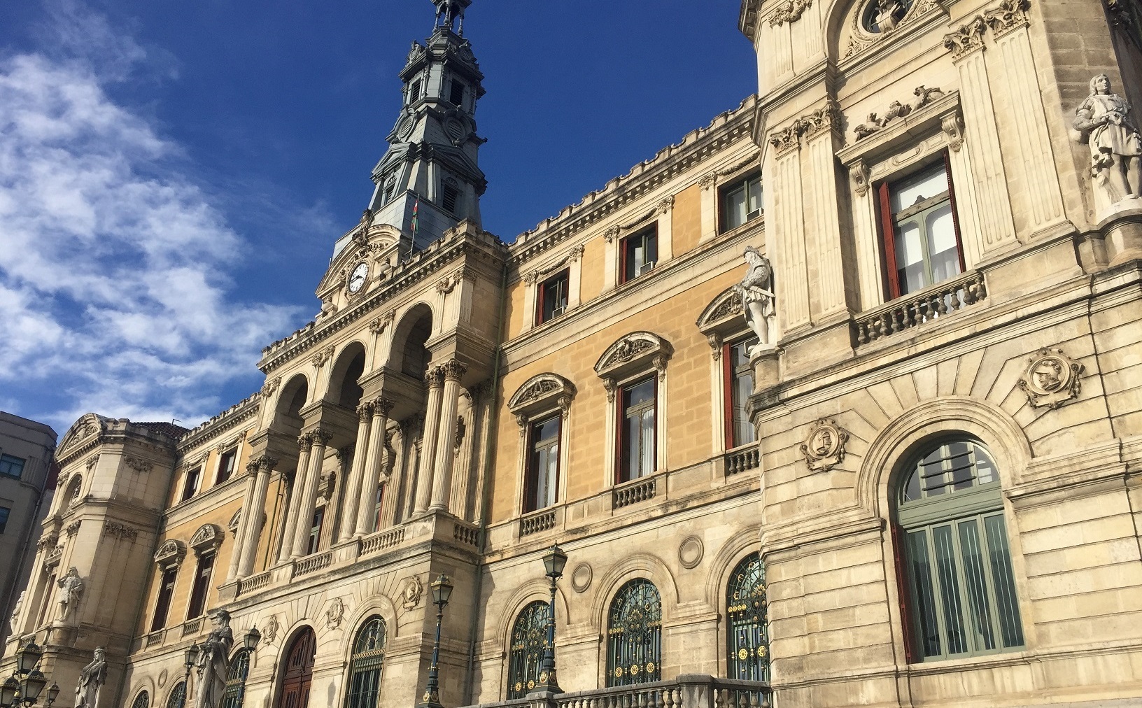 Fachada del Ayuntamiento de Bilbao vista en perspectiva, con un pórtico de arcos y columnas, esculturas de piedra y una torre con reloj bajo un cielo azul.