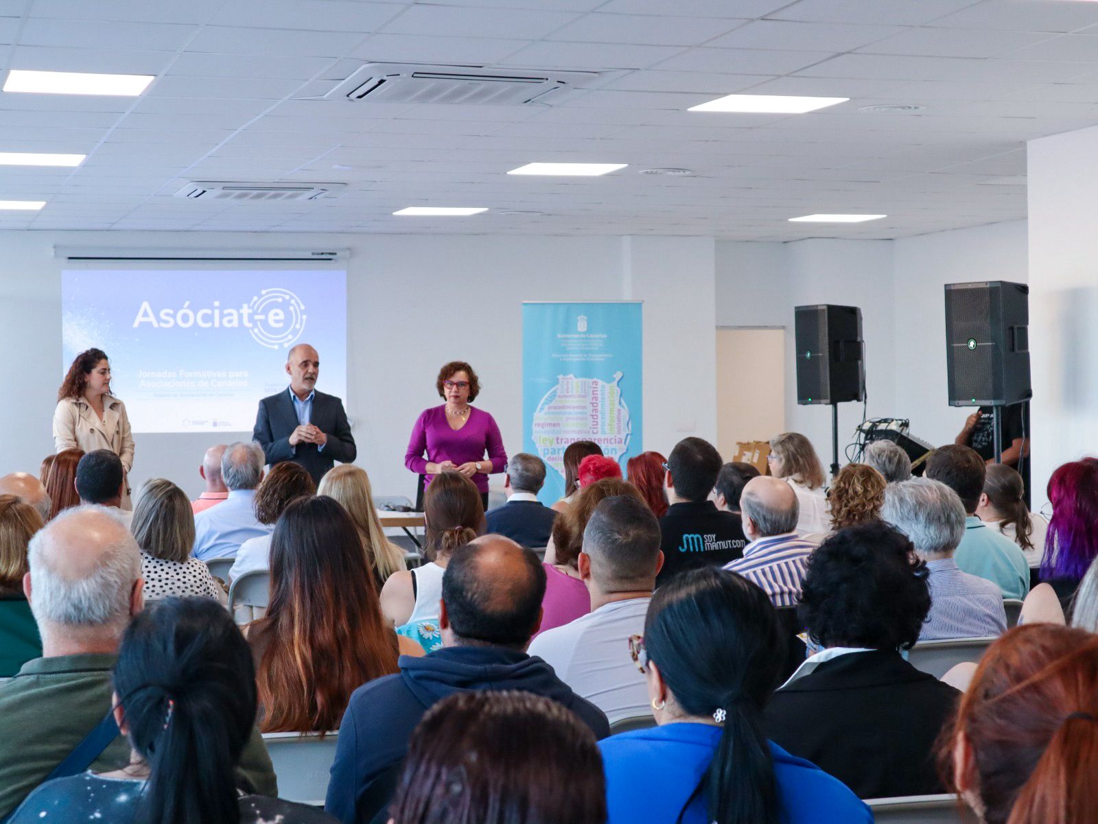 Asistentes sentados en una sala durante una jornada formativa; al fondo, ponentes junto a una pantalla con el logotipo de Asóciat-e.